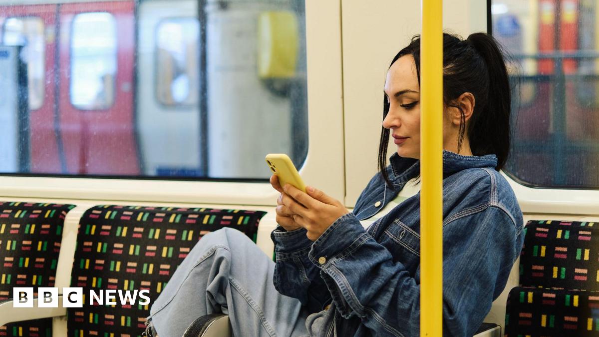 A woman wearing jeans and a denim jacket scrolls on her smartphone whilst on the Tube in London.
