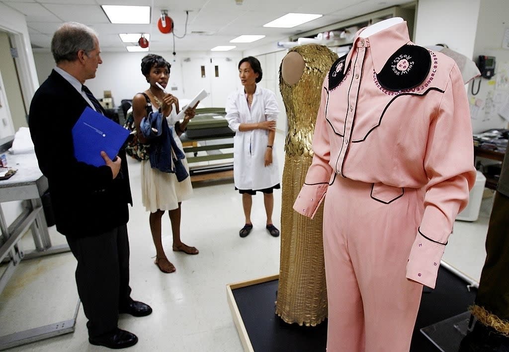 Group discussing costumes in a museum or exhibition space. Pink Western-style outfit with embroidered details is prominently displayed