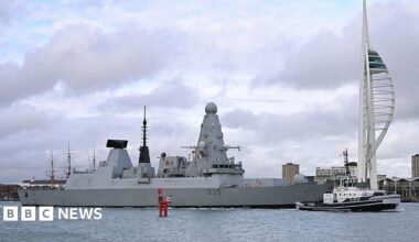 HMS Dragon, a large grey destroyer ship, sits on the water in Portsmouth Harbour, under a cloudy sky.