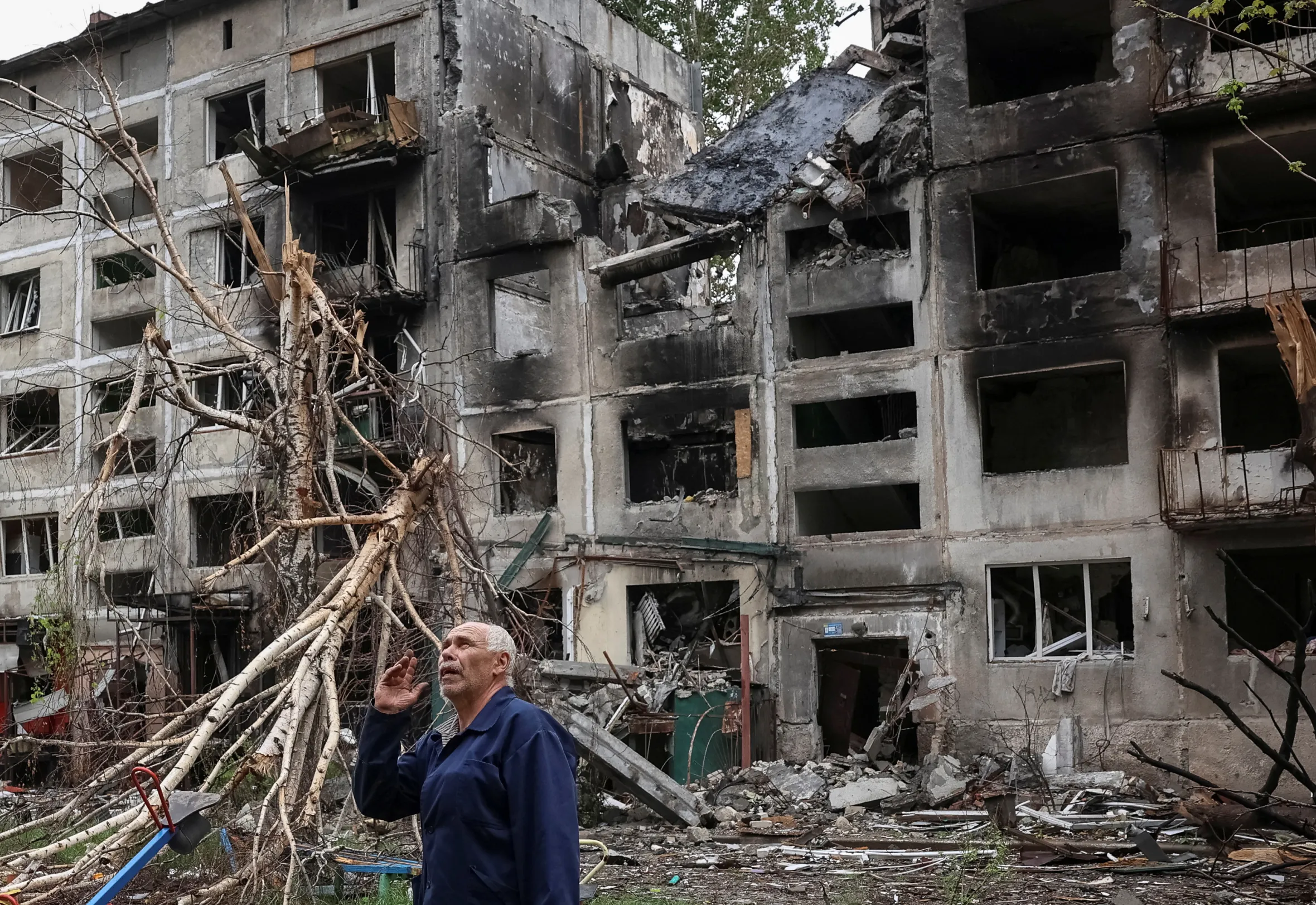 A man gesturing in front of residential buildings damaged by Russian military strikes.