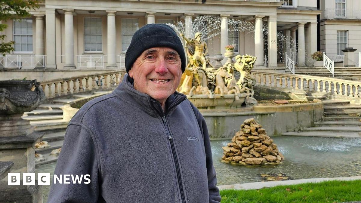 A man in his 70s smiles as he stands in front of an ornate water fountain at the front of a large Regency-style building. He is wearing a black beanie and a grey zip-up fleece. There are steps framing the fountain, which has statues looking over it and a pile of stones in the middle of the water.