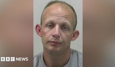 Mugshot of Adam Hall. He is clean shaven and balding with short fair hair, a drooped expression on his face and large ears.