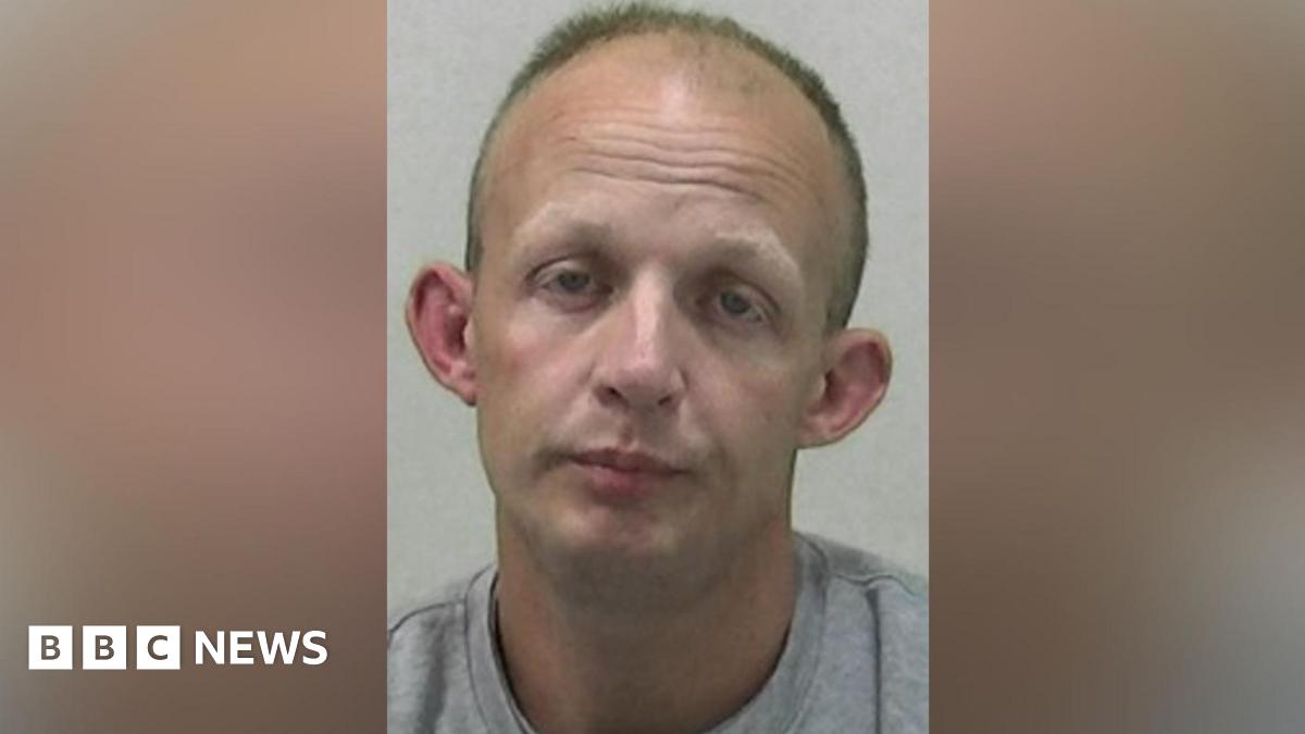 Mugshot of Adam Hall. He is clean shaven and balding with short fair hair, a drooped expression on his face and large ears.