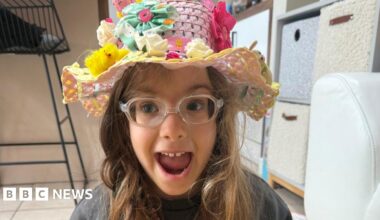 A young girl, with her mouth open, looking excited, wearing clear glasses, and an Easter hat on her head, with chicks, flowers and bright colours on her head. She is by boxes in a room with a chair to the left.