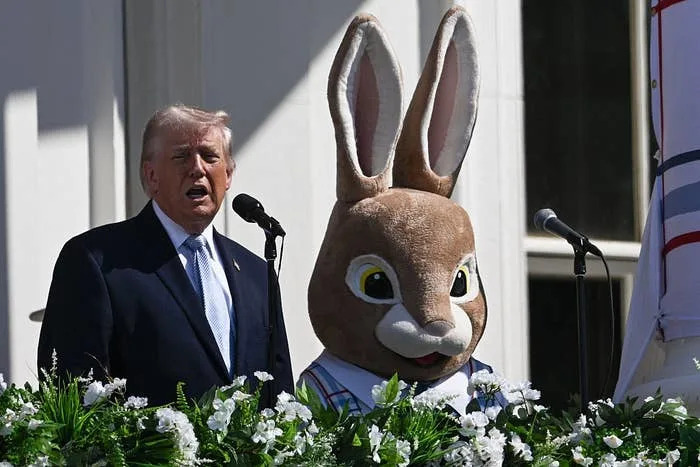 A man in a suit speaks at a podium next to a person in a rabbit costume, on a decorated platform