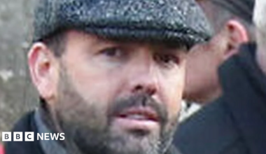 Man looks at camera, he has dark stubble and is wearing a flat cap