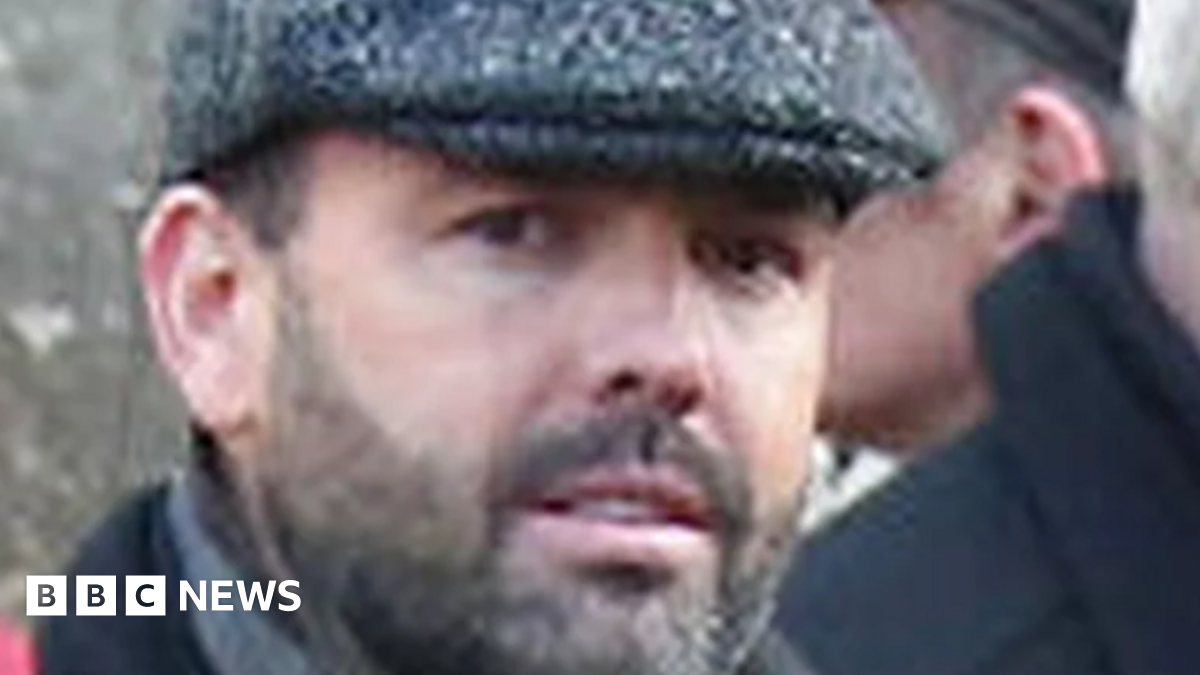 Man looks at camera, he has dark stubble and is wearing a flat cap