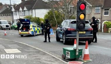 Armed officers can be seen stood next to a black car on a residential road. Another police car is parked to the left and has its boot door open. There is a temporary traffic light in the foreground.