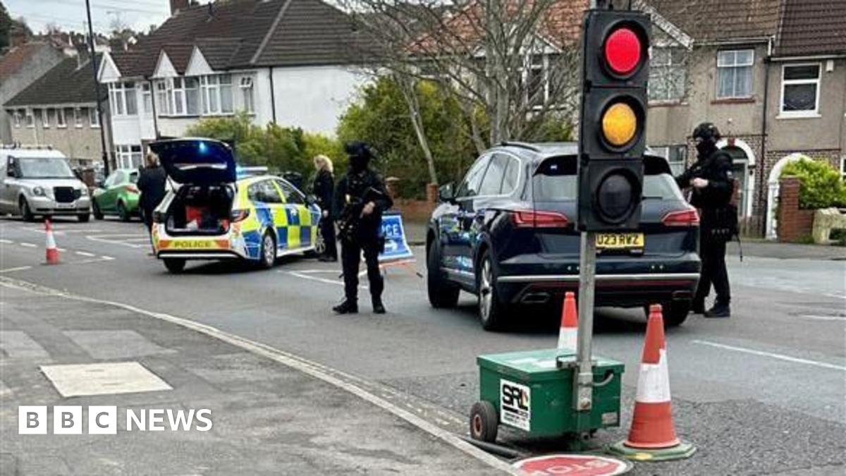 Armed officers can be seen stood next to a black car on a residential road. Another police car is parked to the left and has its boot door open. There is a temporary traffic light in the foreground.