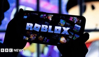 A pair of hands are holding up an iPhone which has the word Roblox in block capital letters on the screen.