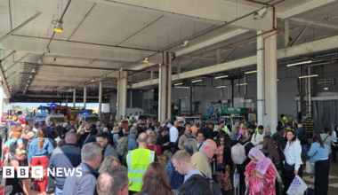 Dozens of passengers stand outside under a roof covering with airport staff in fluorescent bibs. Fire engines can be seen at the rear of the crowds.