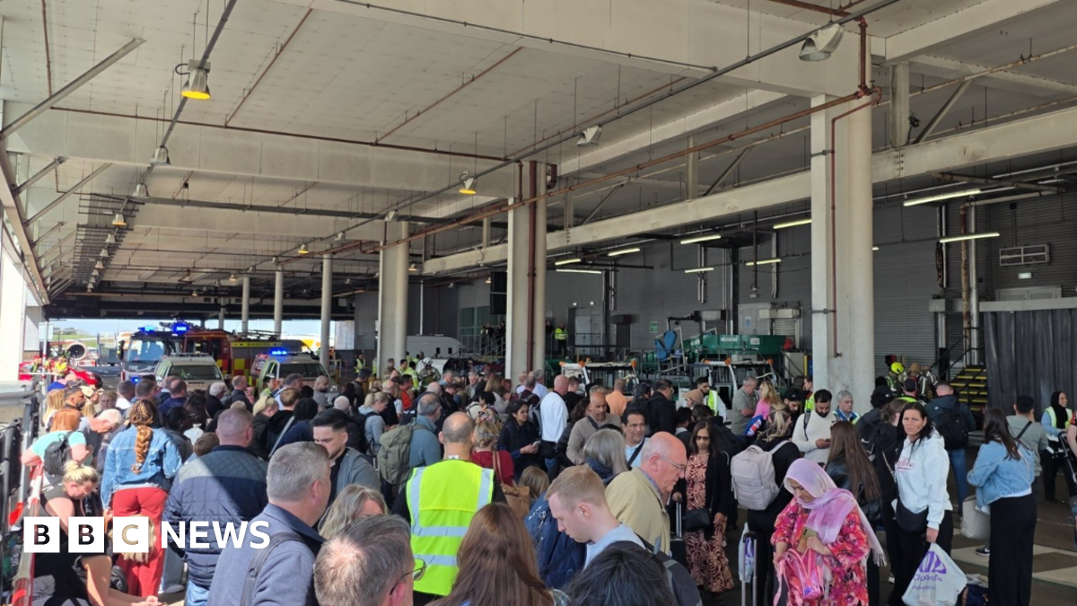 Dozens of passengers stand outside under a roof covering with airport staff in fluorescent bibs. Fire engines can be seen at the rear of the crowds.