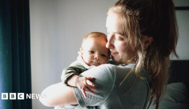 A young mum with light brown hair and wearing a grey T-shirt holds her baby in her arms and looks at him with her head turned to one side, standing in front of a window with daylight streaming through.