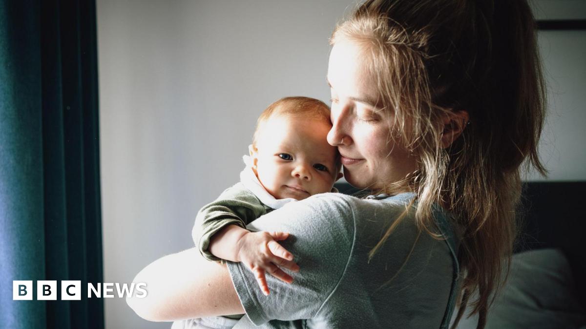 A young mum with light brown hair and wearing a grey T-shirt holds her baby in her arms and looks at him with her head turned to one side, standing in front of a window with daylight streaming through.