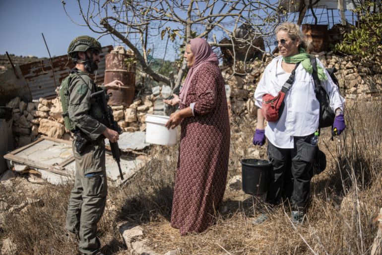 a lady in a pink dress and head scarf holds a white bucket and faces an armed soldier