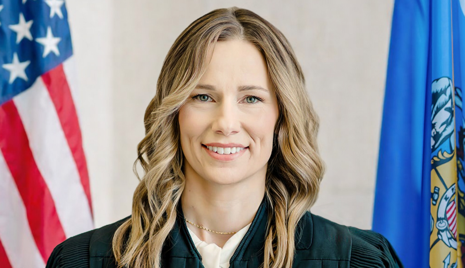 A woman in a judicial robe stands in front of American and Wisconsin state flags, smiling at the camera.