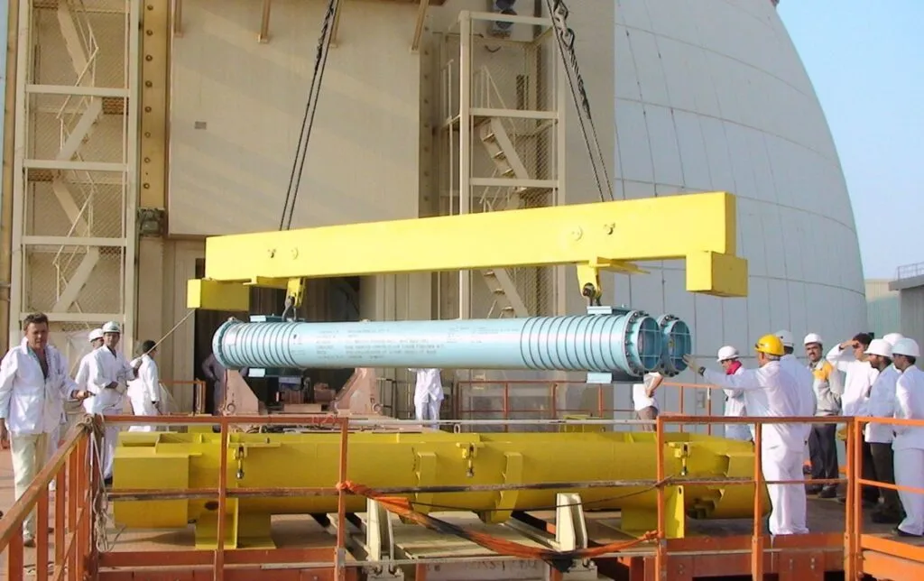 A group of workers in white uniforms and hard hats oversees the lifting of a large blue cylindrical metal component by a yellow crane outside an industrial facility.