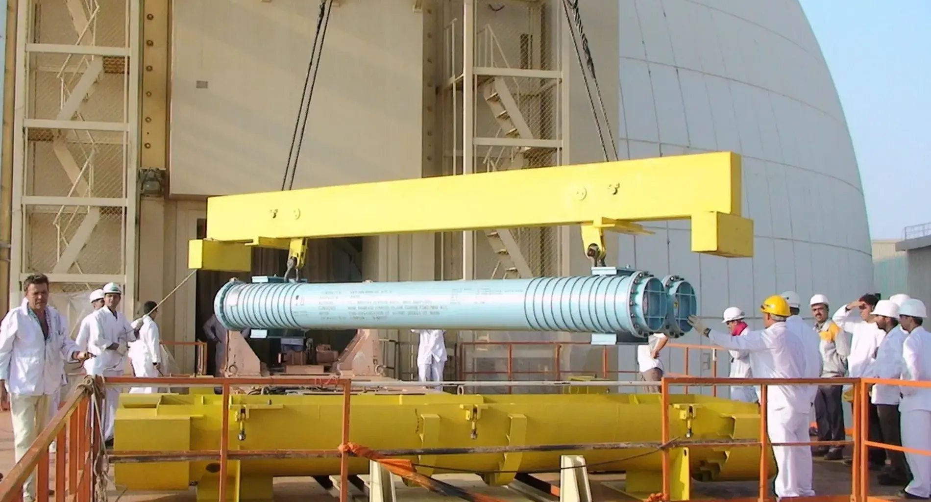 A group of workers in white uniforms and hard hats oversees the lifting of a large blue cylindrical metal component by a yellow crane outside an industrial facility.