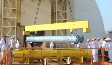 A group of workers in white uniforms and hard hats oversees the lifting of a large blue cylindrical metal component by a yellow crane outside an industrial facility.
