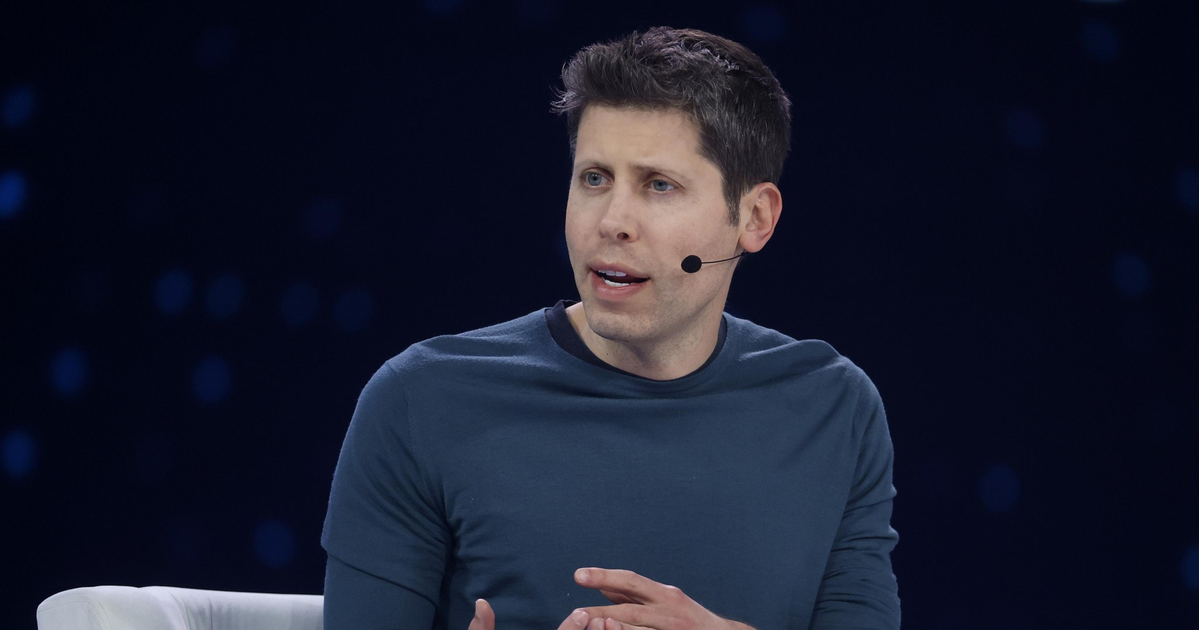 A man wearing a dark blue long-sleeve shirt sits in a white chair, speaking with hand gestures and wearing a headset microphone.