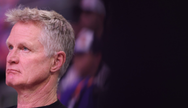 A middle-aged man with short gray hair and a serious expression looks intently to his left, wearing a black shirt against a dark blurred background.