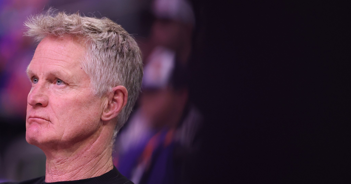 A middle-aged man with short gray hair and a serious expression looks intently to his left, wearing a black shirt against a dark blurred background.
