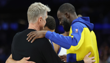 Three men in a group hug, with one wearing a yellow and blue NBA jacket, one in a black shirt, and the third in a white basketball uniform.