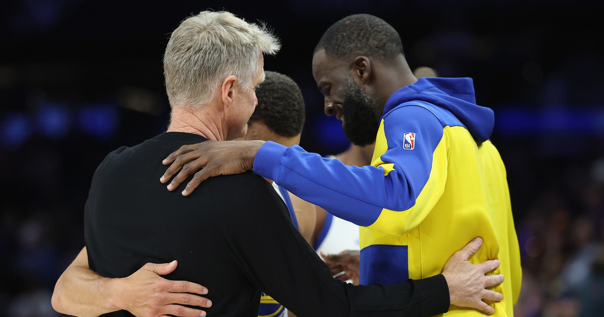 Three men in a group hug, with one wearing a yellow and blue NBA jacket, one in a black shirt, and the third in a white basketball uniform.