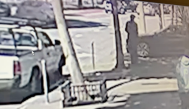 A person stands on a sidewalk near a parked pickup truck and a car, with trees and street signs visible nearby.