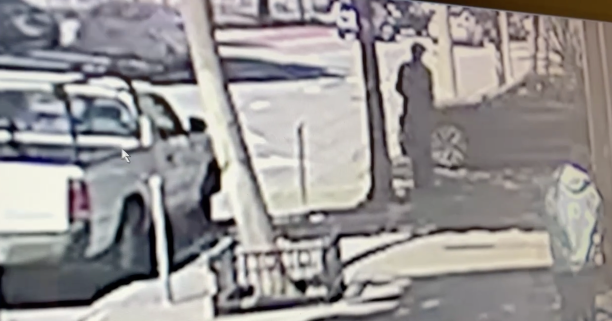 A person stands on a sidewalk near a parked pickup truck and a car, with trees and street signs visible nearby.