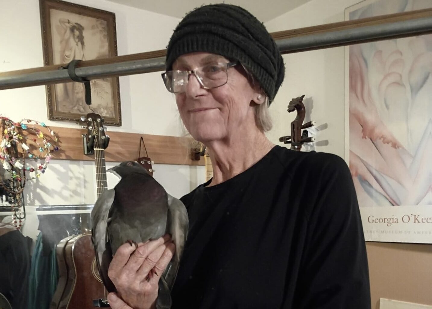 An older person wearing glasses and a knit hat holds a pigeon indoors, with guitars and art posters visible in the background.