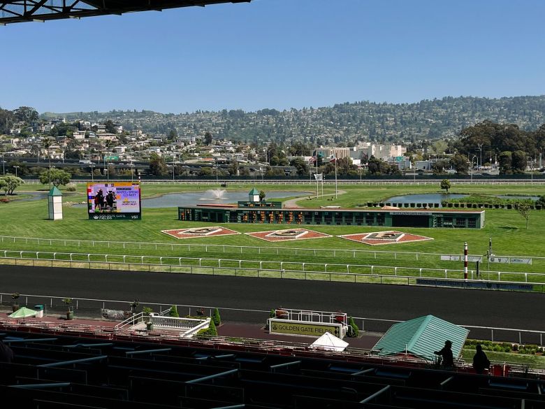 A view of a horse racing track from a grandstand seat, looking out onto a green infield and byond that to the East Bay Hills