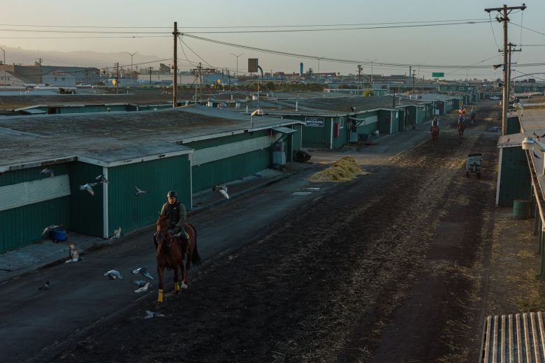 People ride horses along a dirt path, past low-slung green buildings.