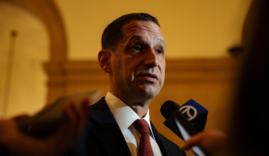 A man in a suit and red tie speaks into microphones, with a golden arch and warm lighting in the background.