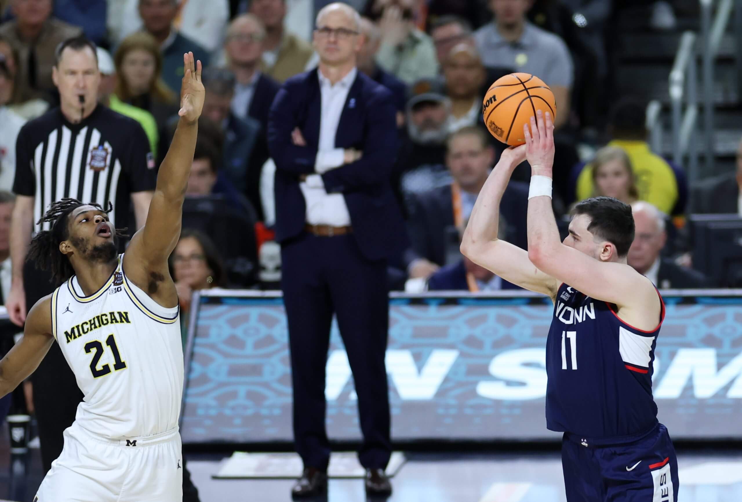 Alex Karaban raises the ball over his head to shoot with a Michigan player jumping to defend and Dan Hurley in the background