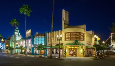 A street scene at night with art deco buildings, illuminated storefronts, and palm trees under a clear sky evokes the vibrant charm of Walt Disney World. Amid the glow, a purple Mickey molded Loungefly bag adds a touch of whimsy to the enchanting atmosphere.