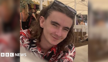 Jamie Collins wearing a black, red and white patterned shirt and sunglasses on his head. He has shoulder length brown hair and piercings in his ears, eyebrow and nose. He appears to be sitting underneath a white marquee on a sunny day.