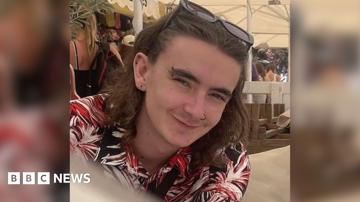 Jamie Collins wearing a black, red and white patterned shirt and sunglasses on his head. He has shoulder length brown hair and piercings in his ears, eyebrow and nose. He appears to be sitting underneath a white marquee on a sunny day.