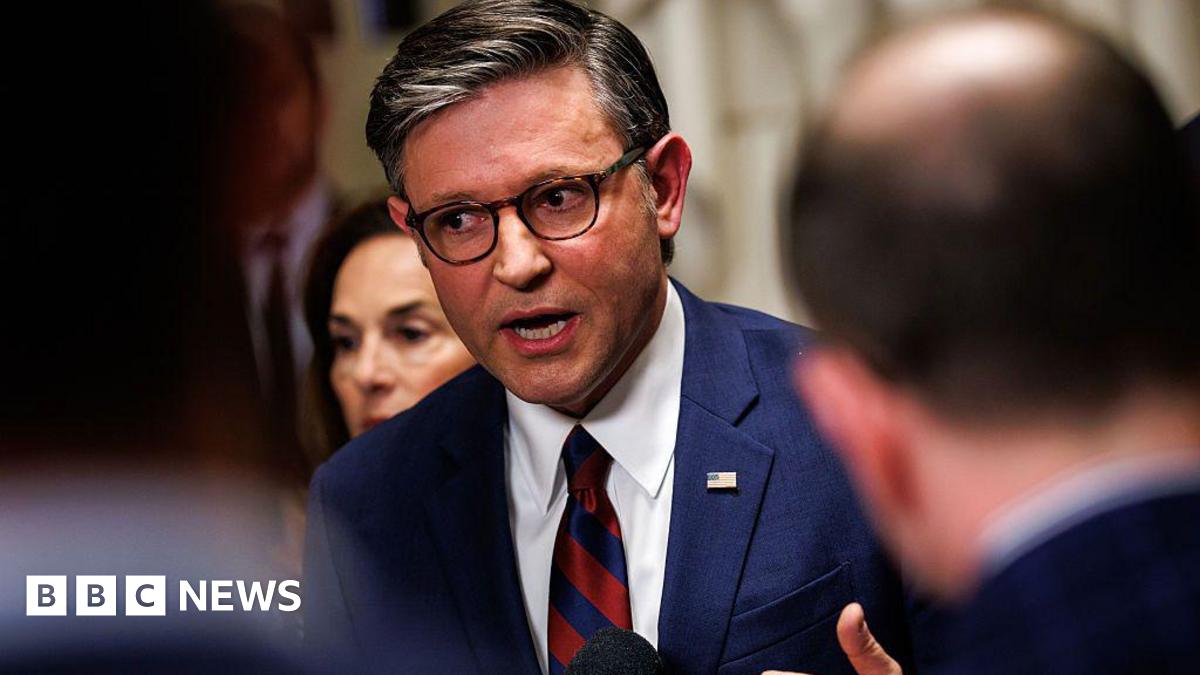 The photo shows four astronauts from NASA’s Artemis II Moon mission standing in a busy indoor sports arena, wearing bright blue flight jackets with mission patches and logos. They are in the foreground, smiling and cheering, with one woman in the centre clasping her hands and two of the men raising their fists in the air in celebration. The crowd behind them is slightly out of focus but fills the stands, suggesting a major sporting event. The lighting is bright and even, and the mood is joyful, energetic and triumphant.