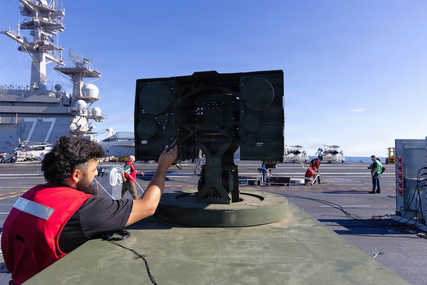 A man sets up a laser system on the deck of an aircraft carrier ship