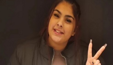 Aaliyah has dark hair in a ponytail, brown eyes and is smiling at the camera. She is wearing a dusty pink t-shirt with a black jacket over the top, she is holding up a peace sign with her fingers.