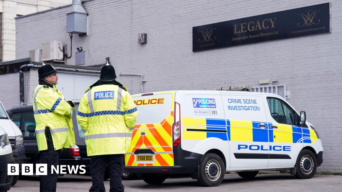 Two police officers wearing yellow jackets and black helmets stand outside a white bricked building. A crime scene investigation police van is parked underneath a black sign that reads Legacy Independent Funeral Directors