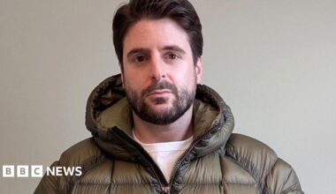 Alex Grossomanides has brown hair, brown eyes and a beard. He is wearing an olive green puffer coat and looking at the camera while standing in front of a white wall indoors.
