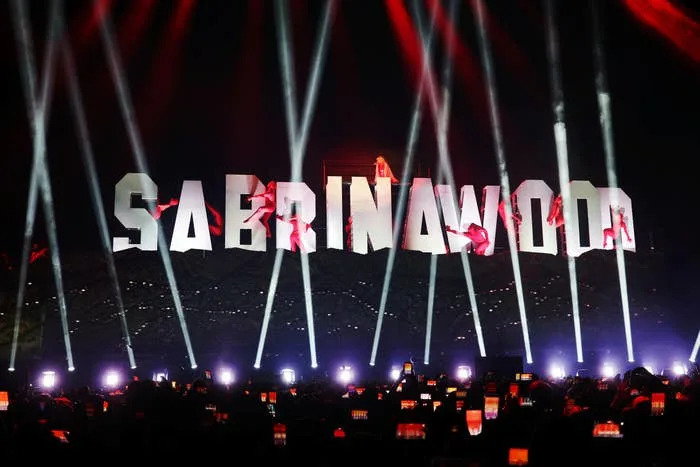Concert stage with large "SABRINAWOOD" letters and energetic light beams. Performers interact with the letters while an audience captures the moment