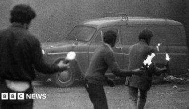 Hilary Benn has short grey hair, round metal glasses, a patterned coat, white shirt and red tie.