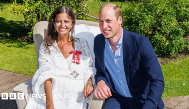 Prince William in a blue suit and shirt crouched next to Dame Deborah James, in a white dress with a medal pinned to it.