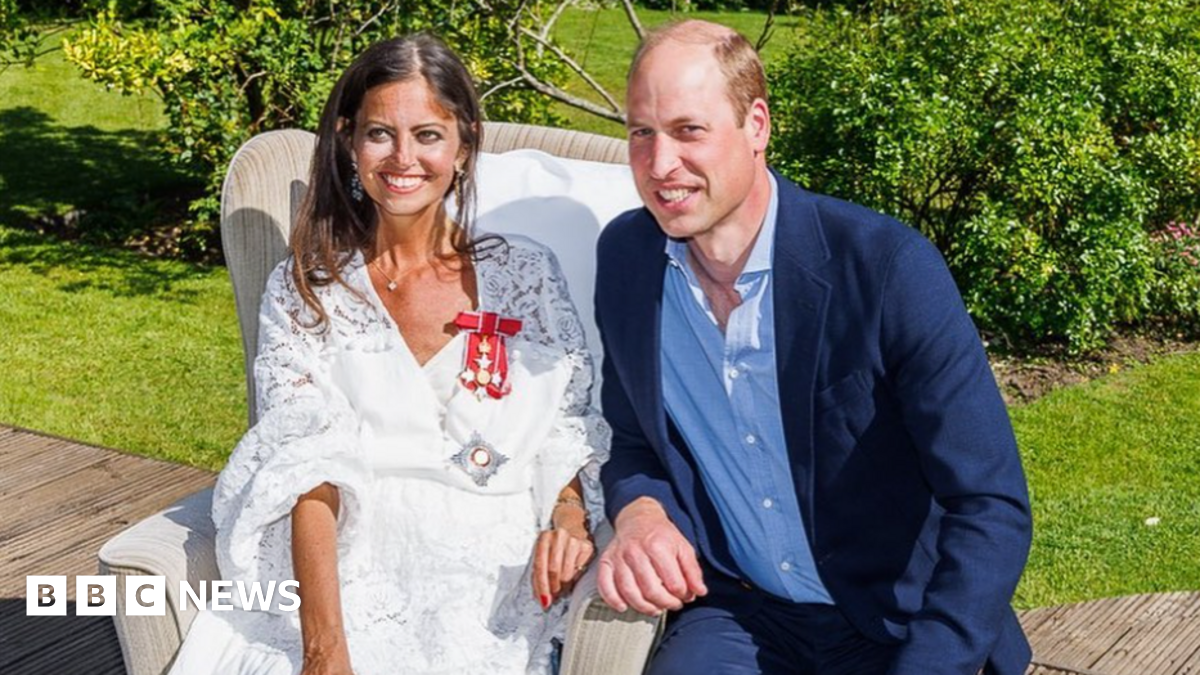 Prince William in a blue suit and shirt crouched next to Dame Deborah James, in a white dress with a medal pinned to it.