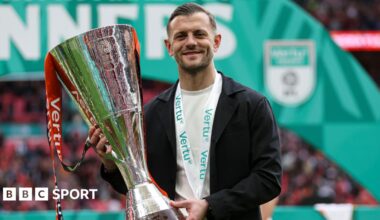 Jack Wilshere, wearing a black blazer and white shirt with a medal around his neck, smiles as he holds up the Vertu Trophy following Luton's 3-1 over Stockport at Wembley