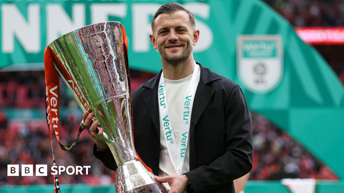 Jack Wilshere, wearing a black blazer and white shirt with a medal around his neck, smiles as he holds up the Vertu Trophy following Luton's 3-1 over Stockport at Wembley