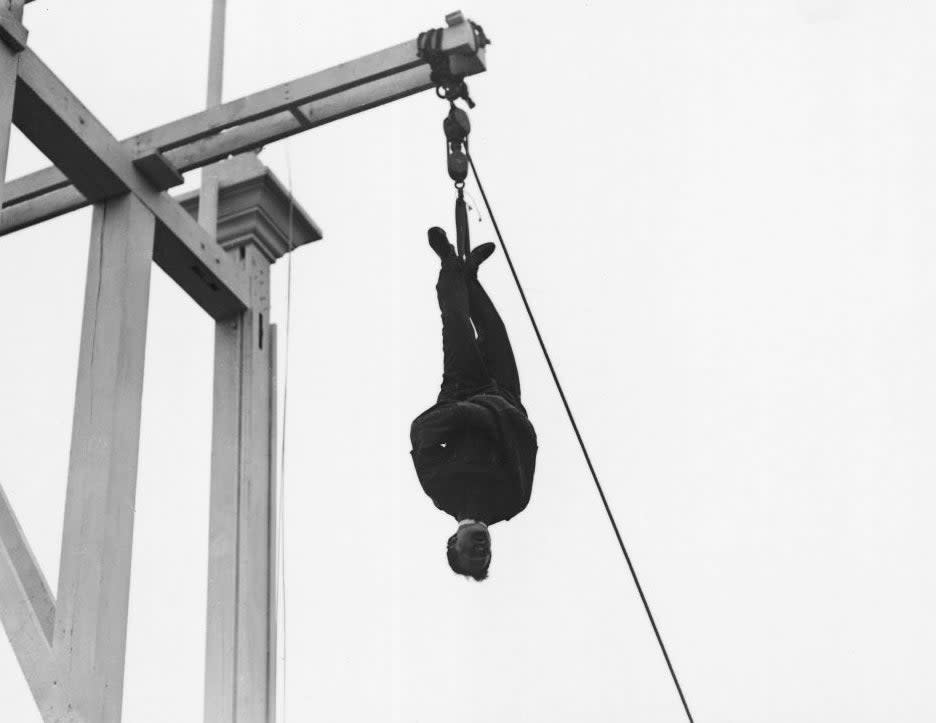 A person suspended upside down from a high structure, demonstrating a physical stunt. The setting appears industrial